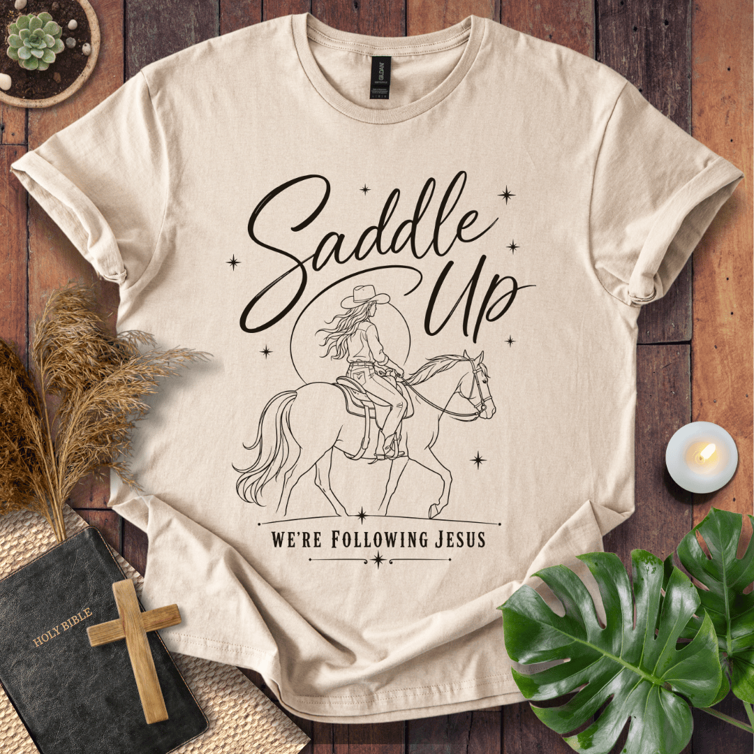 "Saddle Up, We're Following Jesus" shirt with a cowgirl riding a horse under a moonlit sky, displayed on a wooden background.