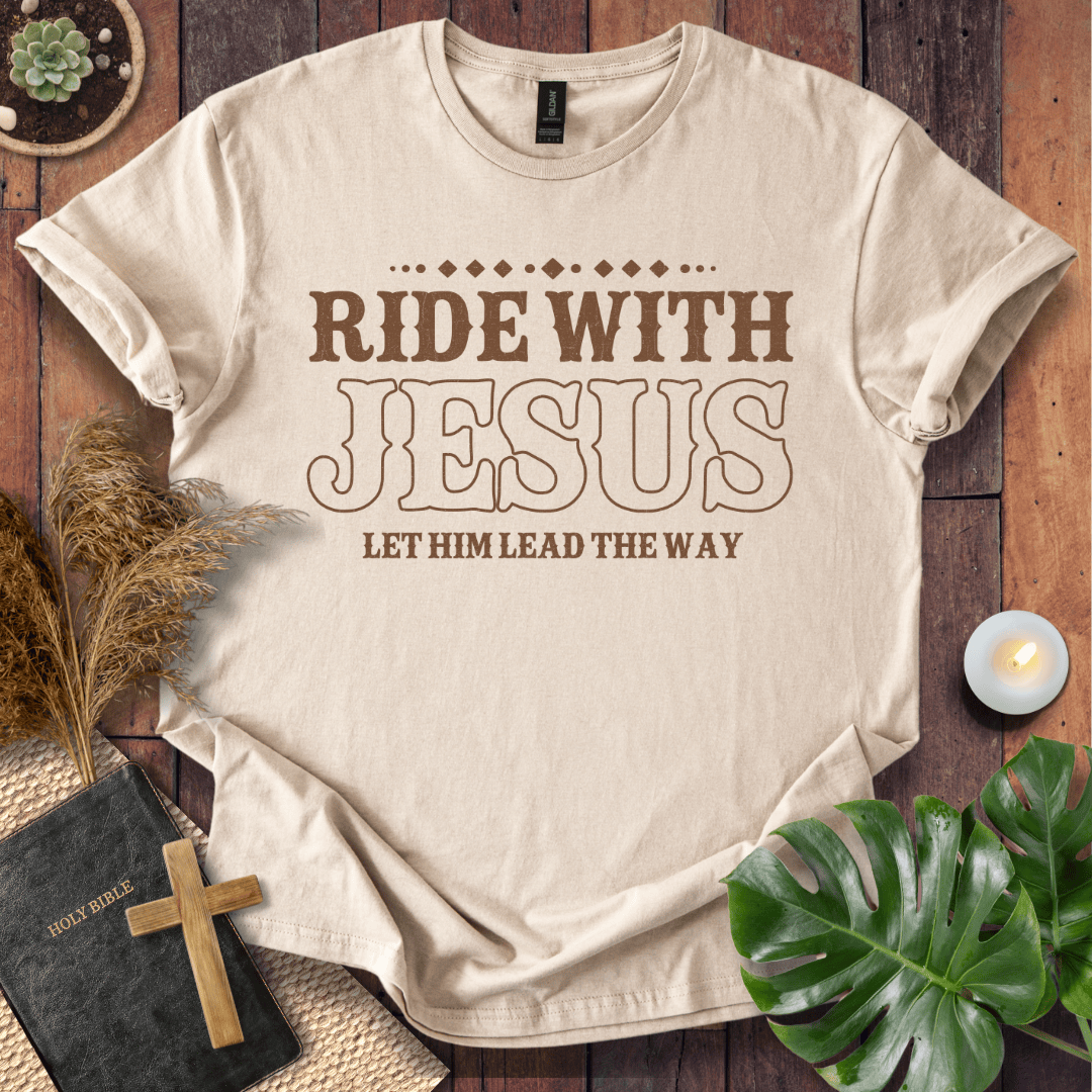 Ride With Jesus Western graphic tee on a wooden background with a Bible, cross, and green leaves.