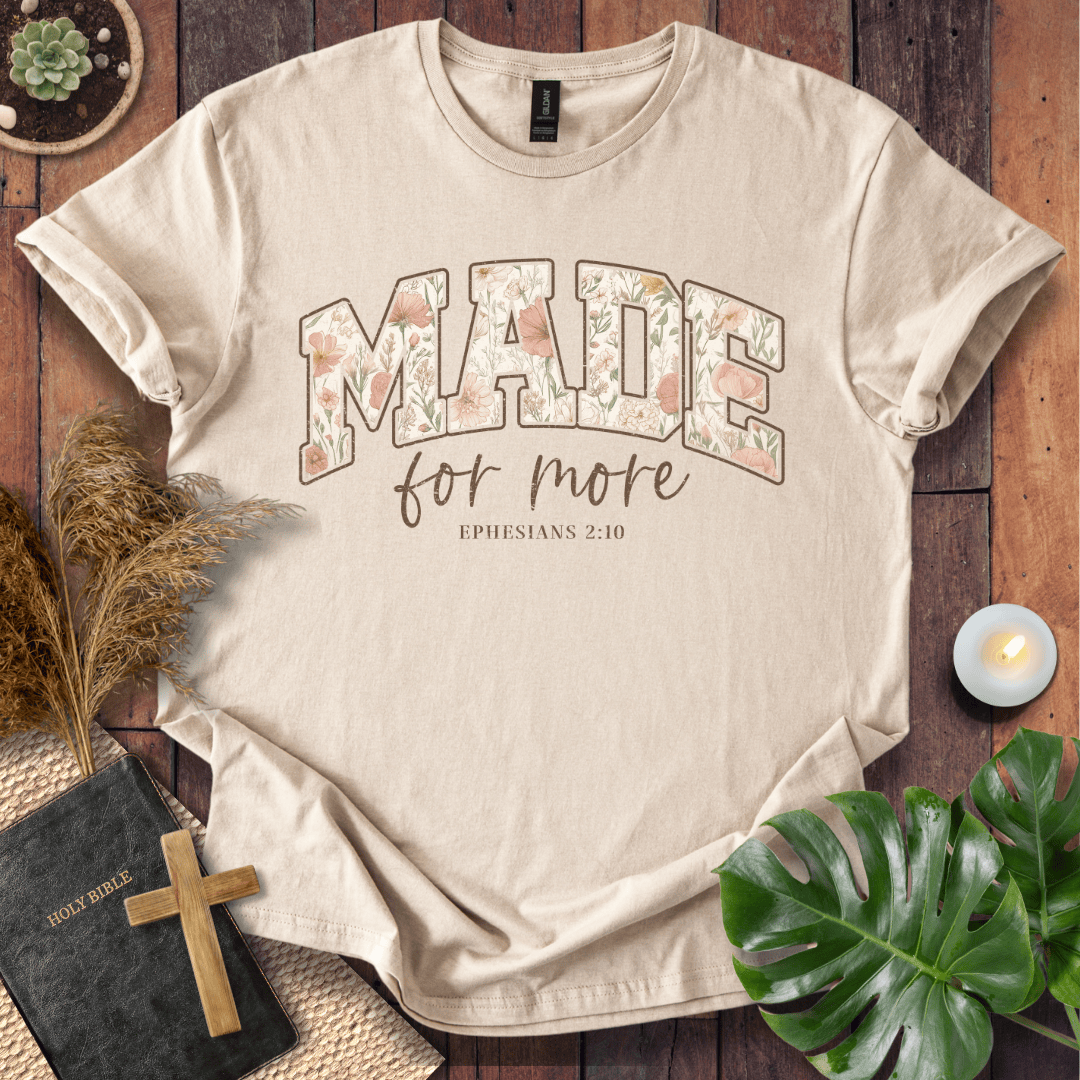 A beige short-sleeve graphic T-shirt laid flat on a rustic wooden surface, surrounded by decorative props including a small cross, a Bible, dried grass, a candle, and green leaves. The shirt has large floral-filled letters reading “MADE” with smaller text below reading “for more” and “EPHESIANS 2:10.