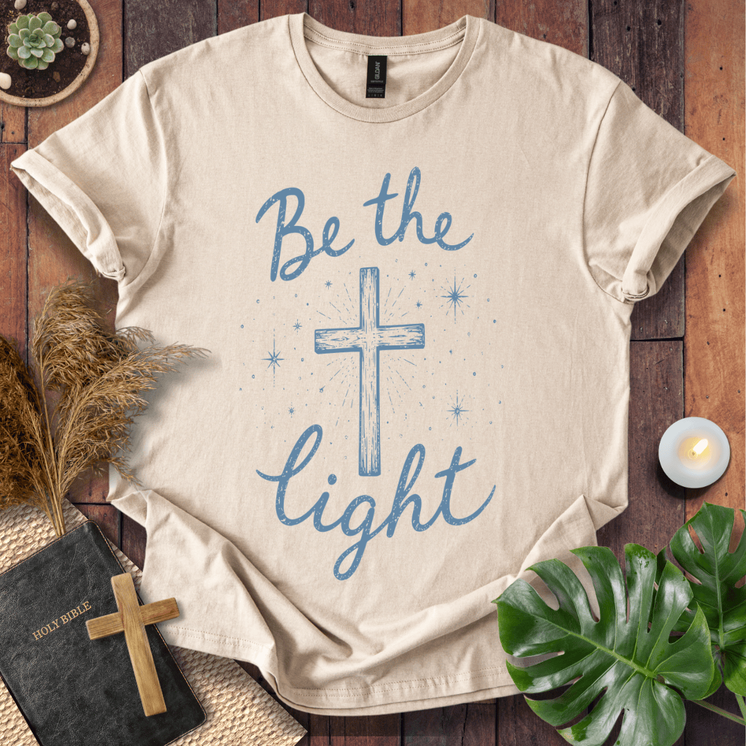 Christian t shirt for women with blue "Be the Light" text, a central cross, and starry accents on a wooden background.