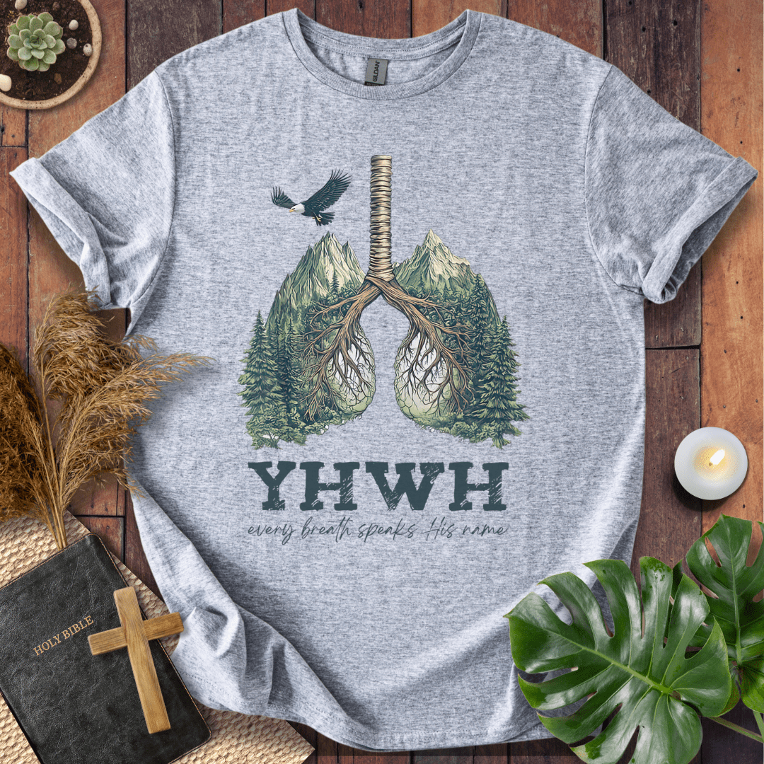 Christian t-shirt with YHWH design, tree-lung illustration, mountains, and “Every Breath Speaks His Name” text, laid flat with Bible and plants.