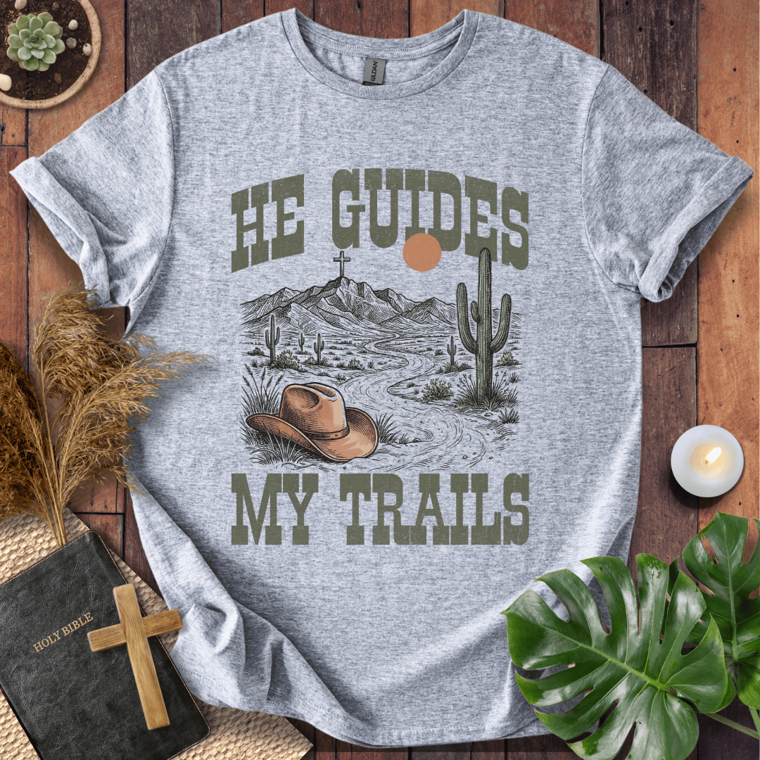 A Christian t-shirt with a desert landscape, cowboy hat, and cross, reading He Guides My Trails.
