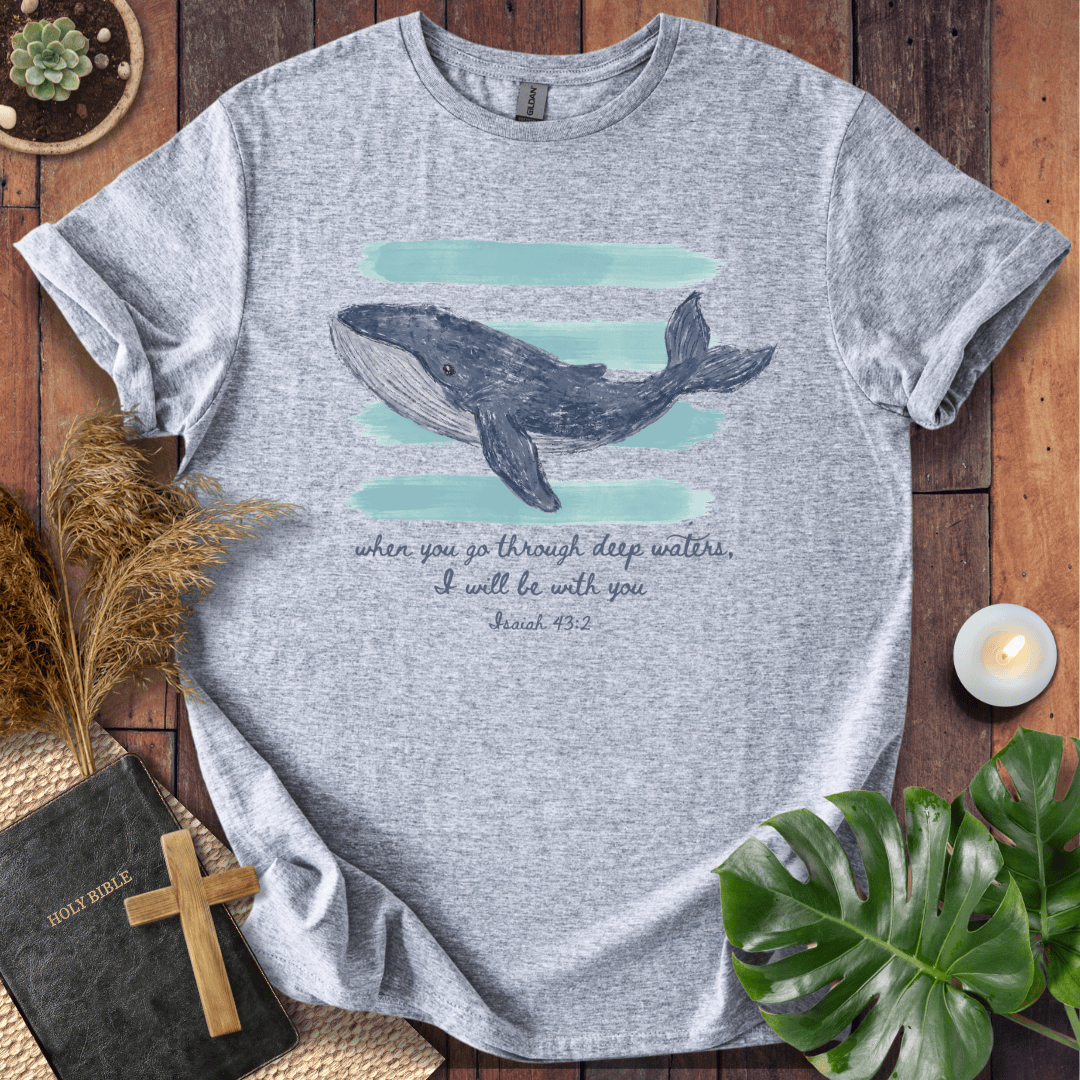 A heather gray short-sleeve T-shirt laid flat on a rustic wood surface, featuring a whale illustration above the printed phrase, 'when you go through deep waters, I will be with you Psalm 48:20.' The shirt is styled with a book, cross, dried grass, candle, and green leaves around it.