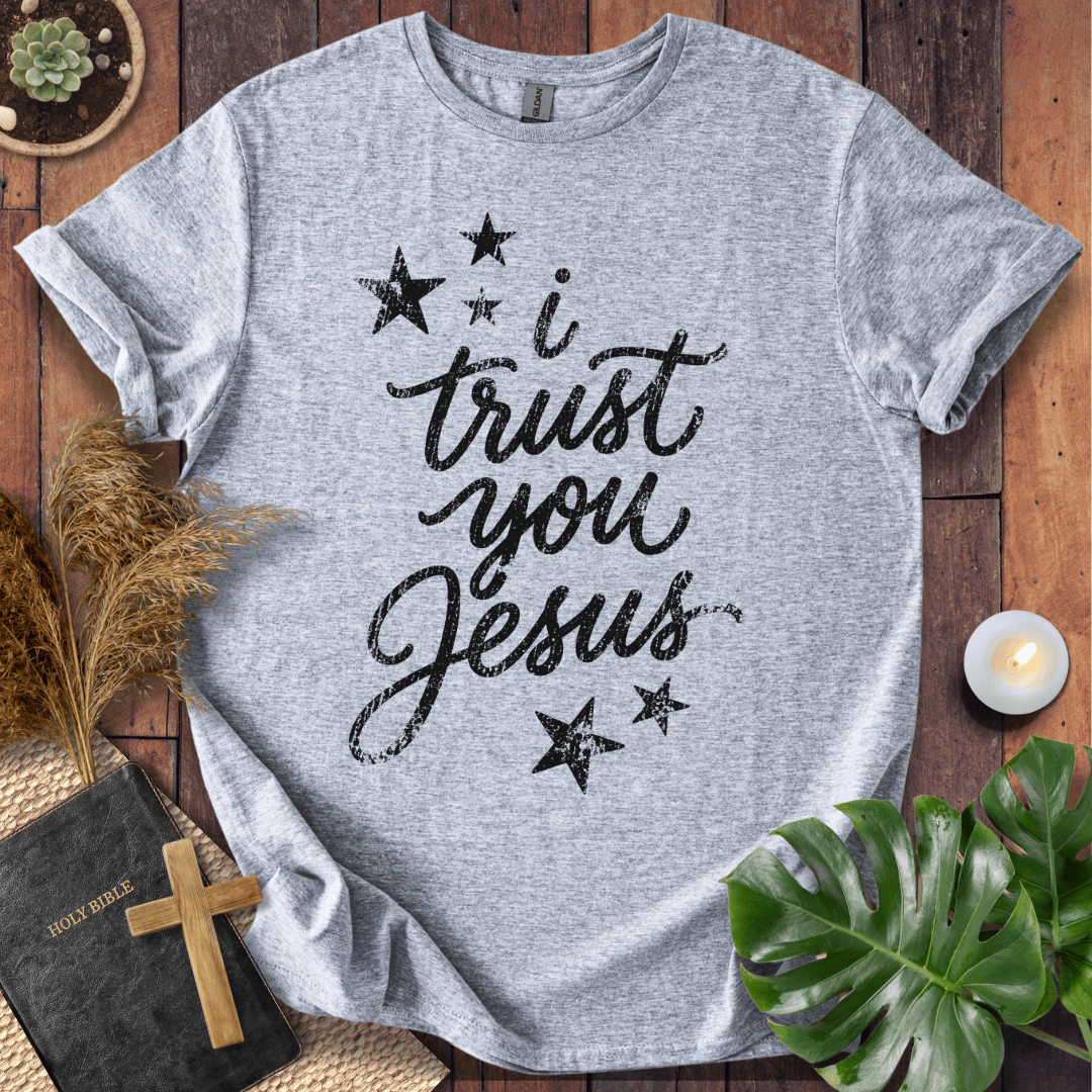 A heather gray short-sleeve T-shirt laid flat on a rustic wooden surface, printed with the phrase 'i trust you Jesus' in black script and surrounded by black stars. The shirt is styled with a Bible, wooden cross, dried grass, green leaves, and a small candle.