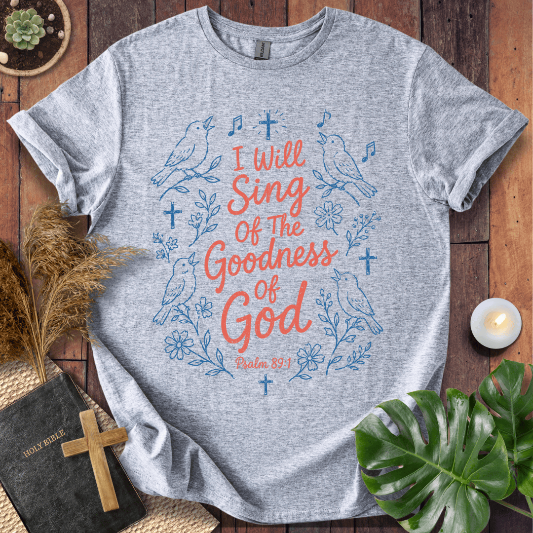 A heather gray graphic t-shirt laid flat on a dark wood background, surrounded by decorative props including a Bible with a wooden cross, dried grass, a small candle, and green leaves. The t-shirt features a blue and coral graphic with birds, flowers, and the text 'I Will Sing Of The Goodness Of God Psalm 89:1'.