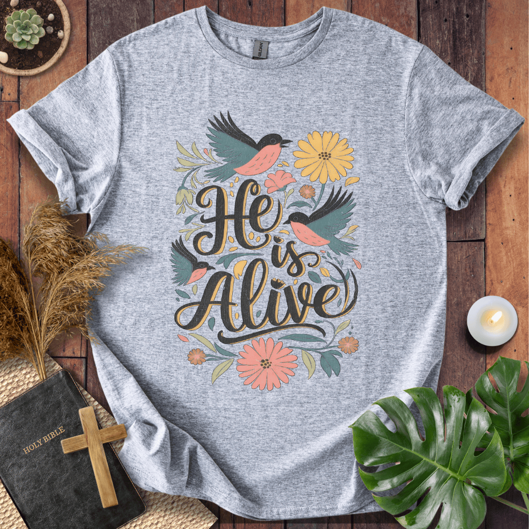 He Is Alive" Christian Easter t shirt for women with birds and flowers on a natural cream shirt, styled on a rustic wood background.