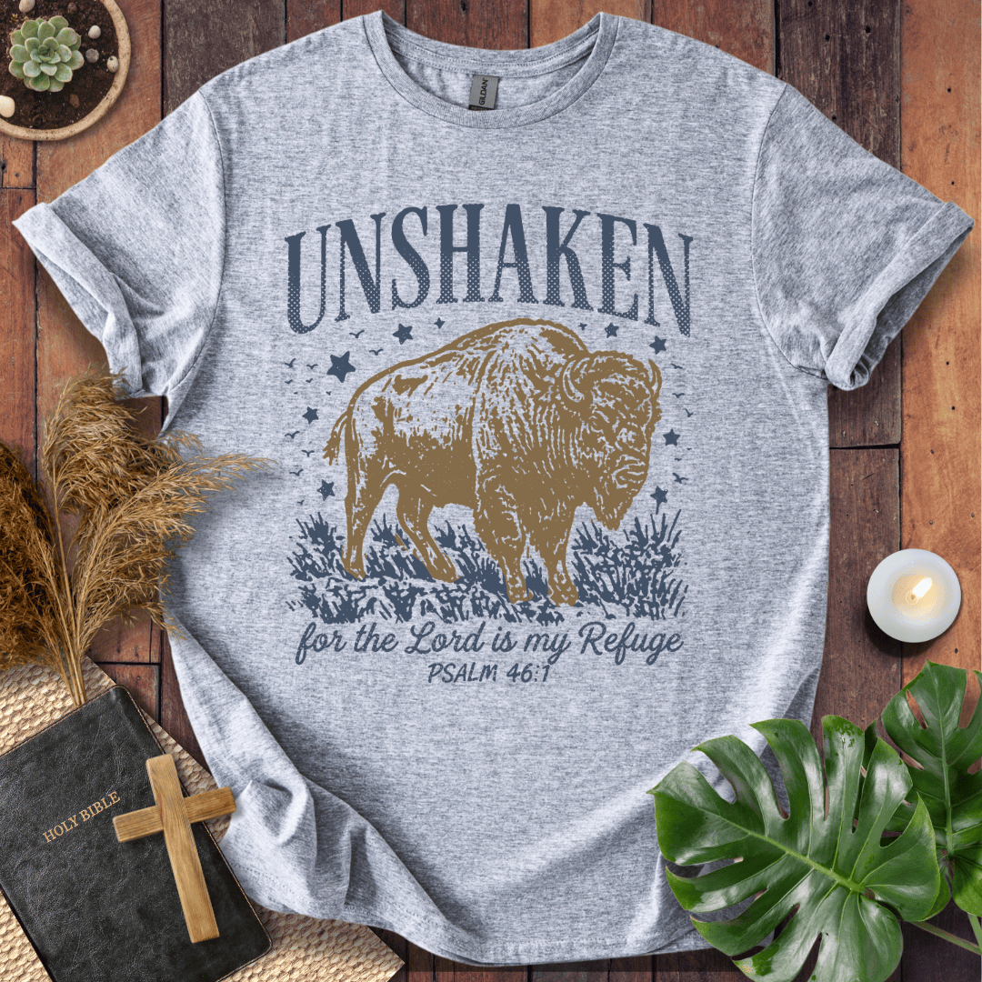 A heather gray graphic t-shirt featuring a brown bison and the text 'UNSHAKEN for the Lord is my Refuge PSALM 46:1' printed on the front. The t-shirt is styled in a flat-lay arrangement on a wooden surface with a Bible, a wooden cross, a small candle, dried grass, and green leaves.