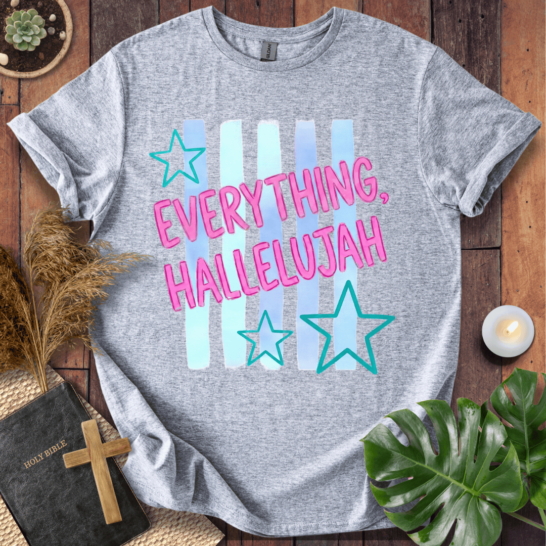A heather gray short-sleeve T-shirt laid flat on a wooden surface, featuring the phrase 'EVERYTHING, HALLELUJAH' in pink outlined letters over pastel vertical stripes and teal star graphics. The shirt is styled with a Bible, cross, pampas grass, candle, succulent, and green leaves around it.