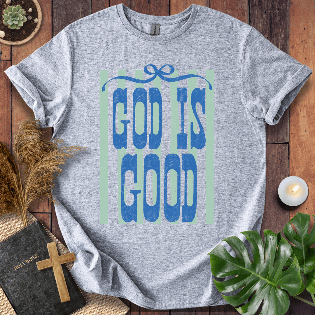 A heather gray short-sleeve T-shirt laid flat on a rustic wooden surface, featuring a large graphic with the words “GOD IS GOOD” in blue letters over a pale green rectangular background. The shirt is styled with a black Bible, a wooden cross, dried grass, a small candle, and green leaves around it.