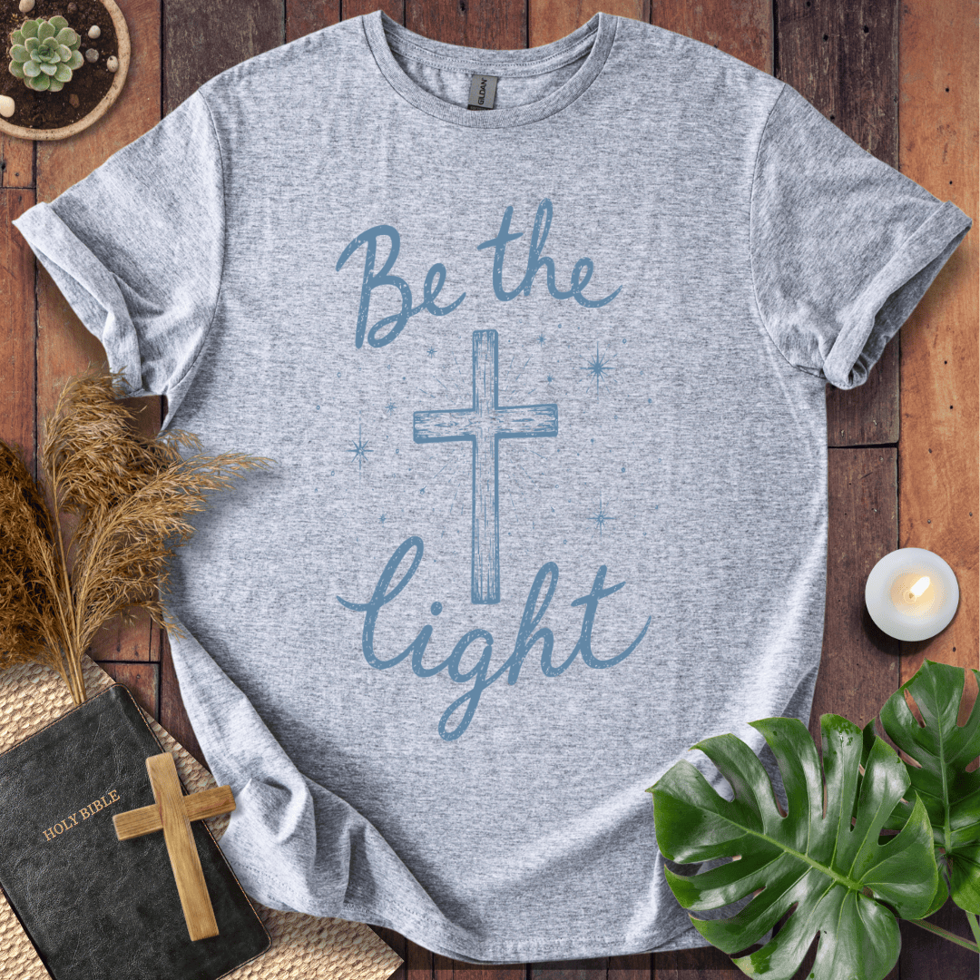 Christian t shirt for women with blue "Be the Light" text, a central cross, and starry accents on a wooden background.