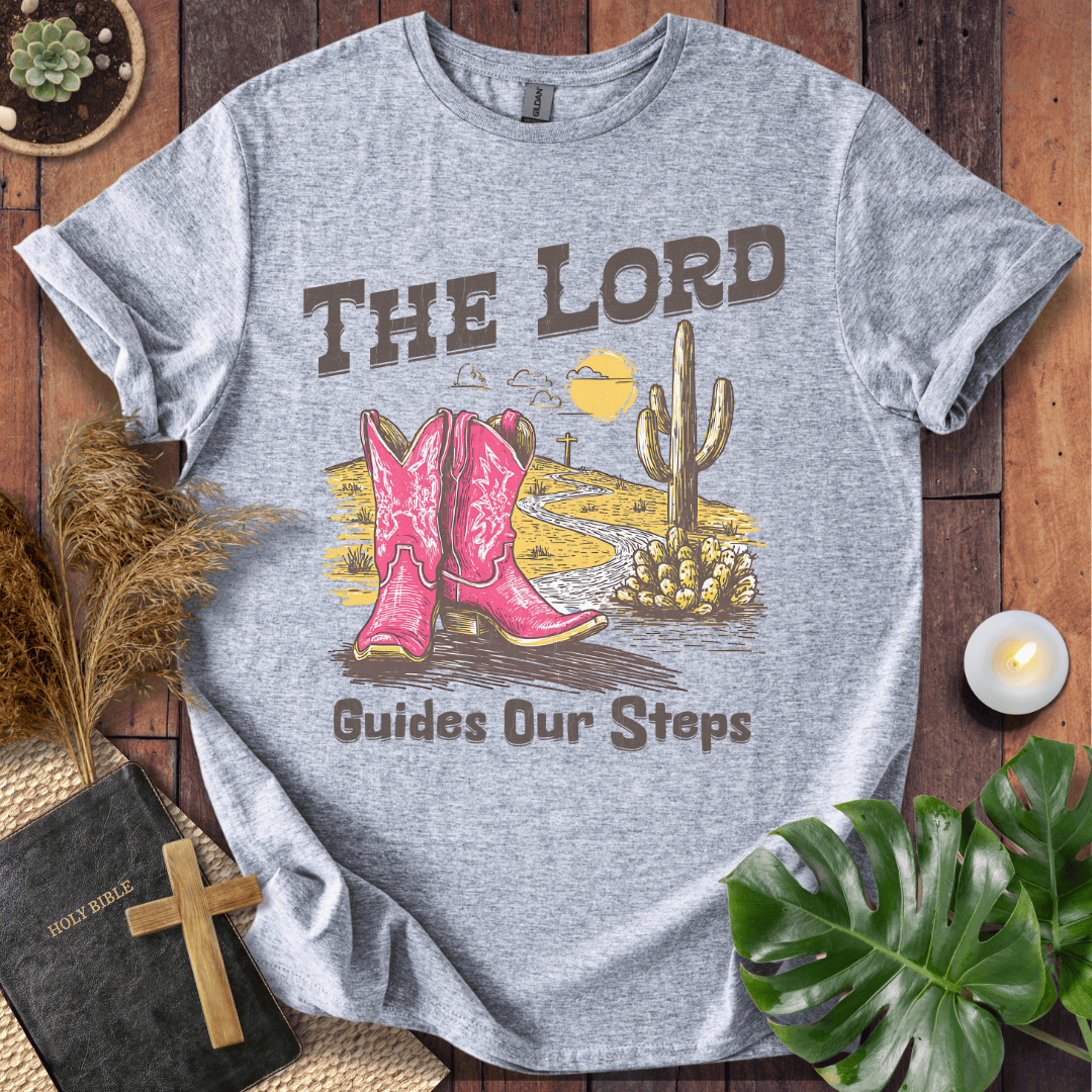 t-shirt with pink cowboy boots and "The Lord Guides Our Steps" text in a desert landscape with a cross and cactus.