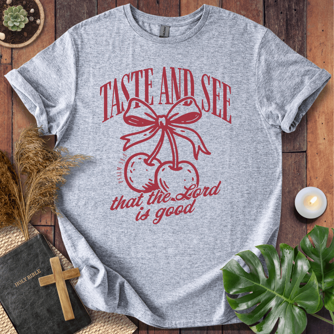 A heather gray short-sleeve t-shirt with a red graphic print featuring cherries under a bow and the text 'TASTE AND SEE that the Lord is good'. The shirt is displayed in a styled flat-lay on a dark wood background with a Bible, wooden cross, pampas grass, a lit candle, and a green monstera leaf.