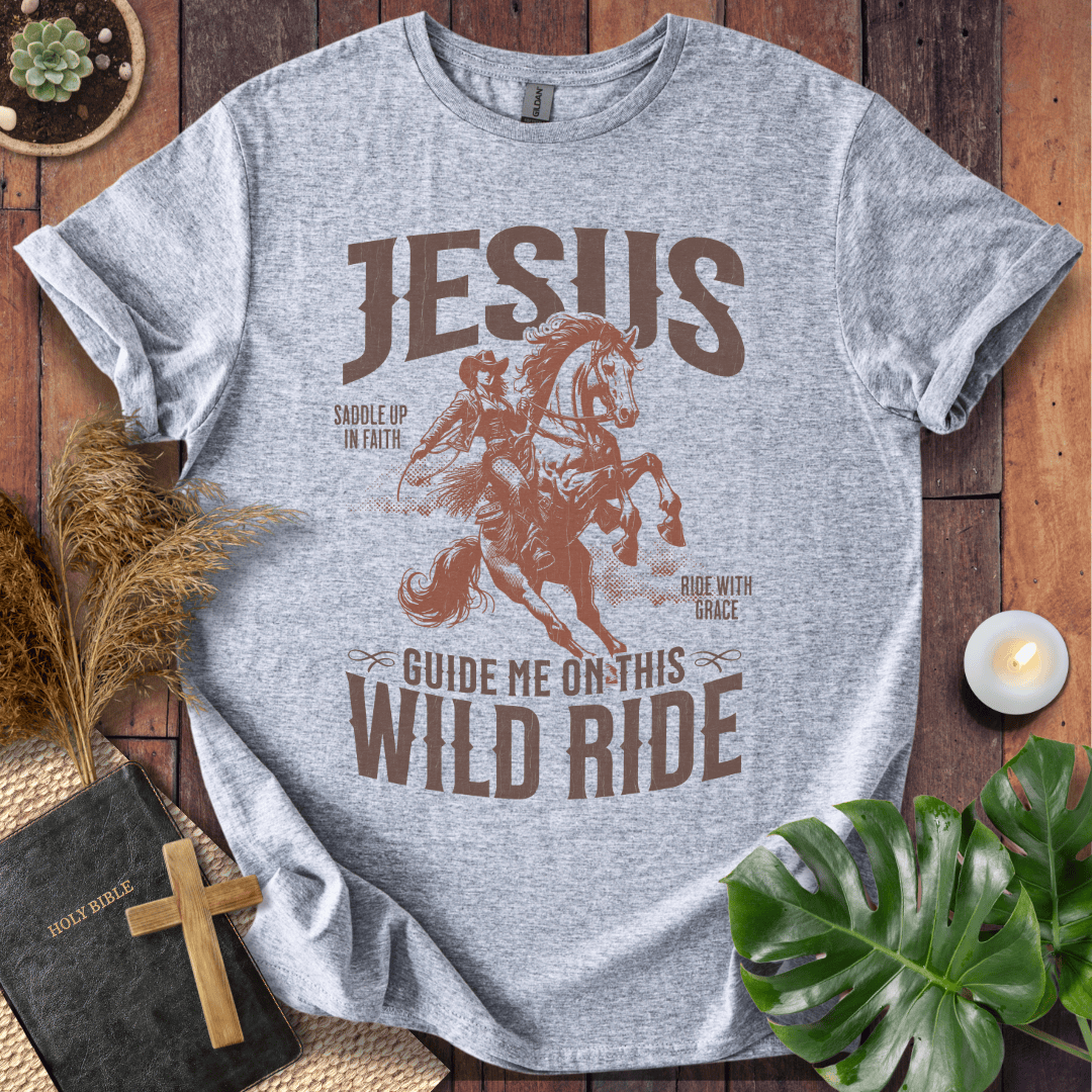 Gray t-shirt with 'Jesus Guide Me on This Wild Ride' graphic on a wooden surface with decor items.