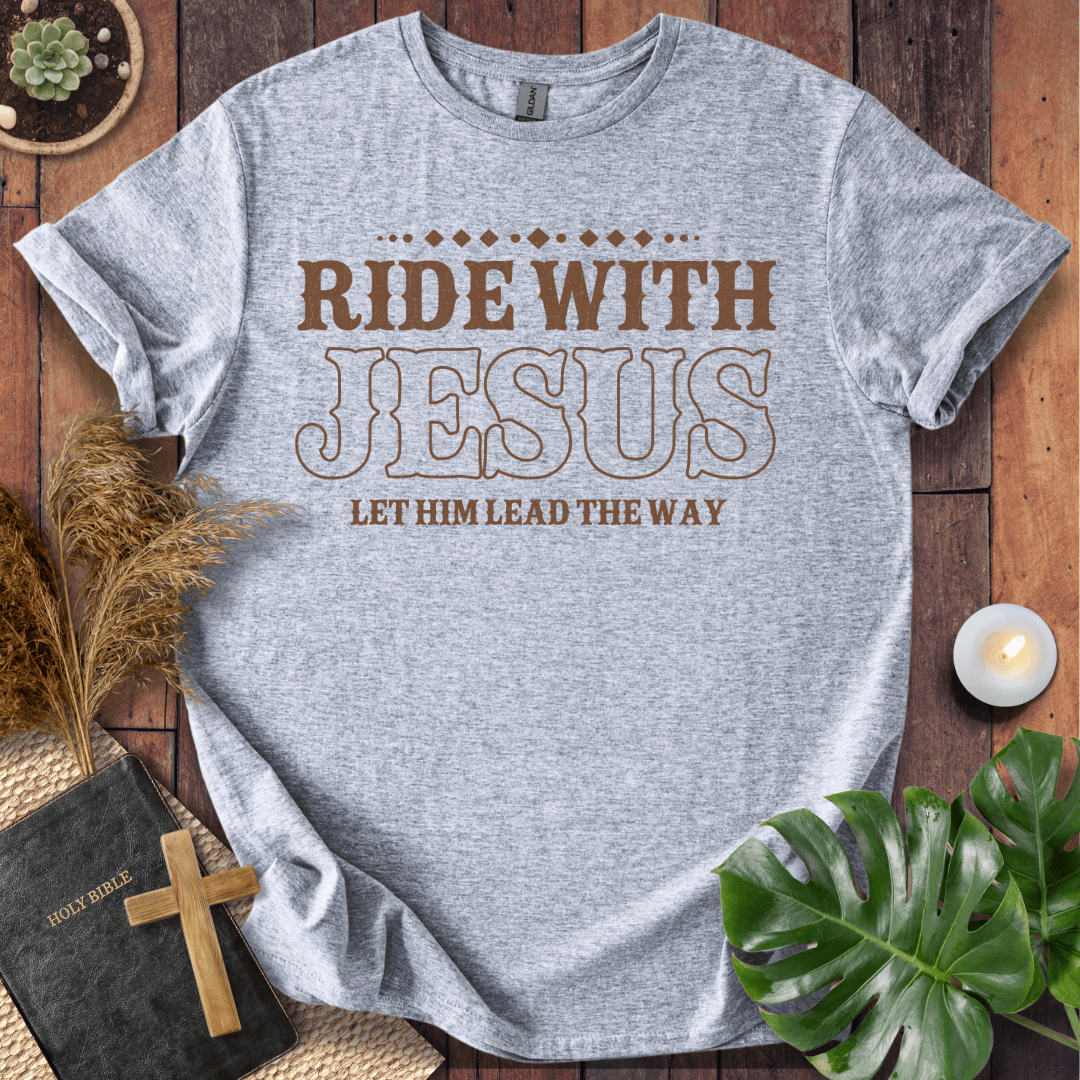Ride With Jesus Western graphic tee on a wooden background with a Bible, cross, and green leaves.
