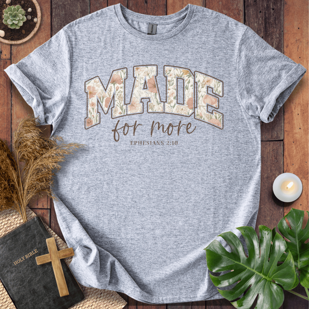 A heather gray graphic t-shirt laid flat on a rustic wooden surface, surrounded by decorative props including a Bible, cross, dried grass, a candle, and green leaves. The shirt features a graphic that reads 'MADE for more' with 'EPHESIANS 3:18' underneath.