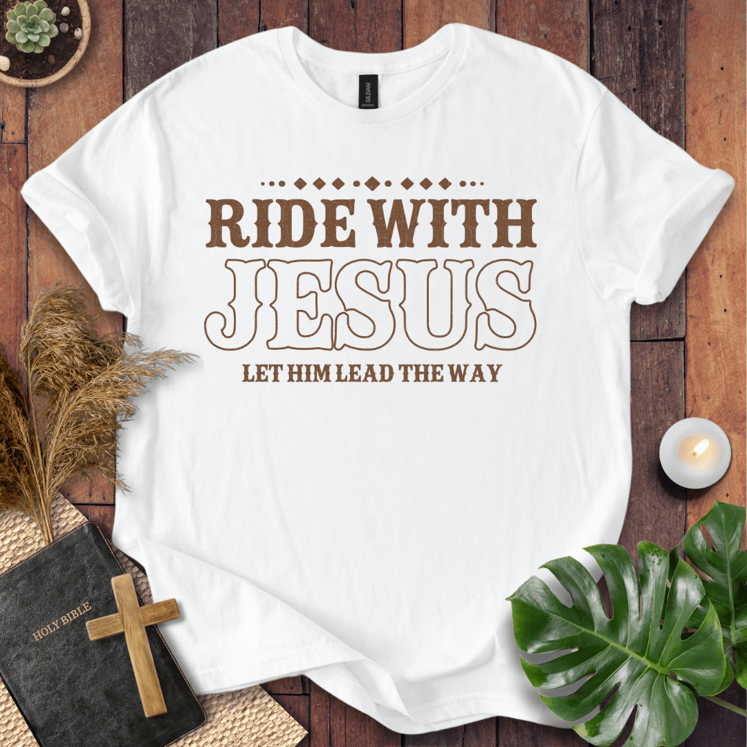 Ride With Jesus Western graphic tee on a wooden background with a Bible, cross, and green leaves.