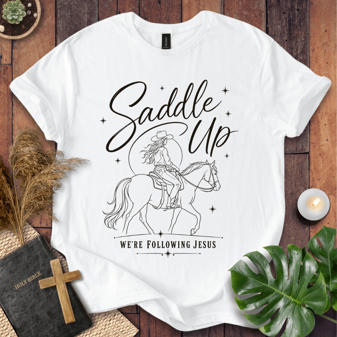 "Saddle Up, We're Following Jesus" shirt with a cowgirl riding a horse under a moonlit sky, displayed on a wooden background.