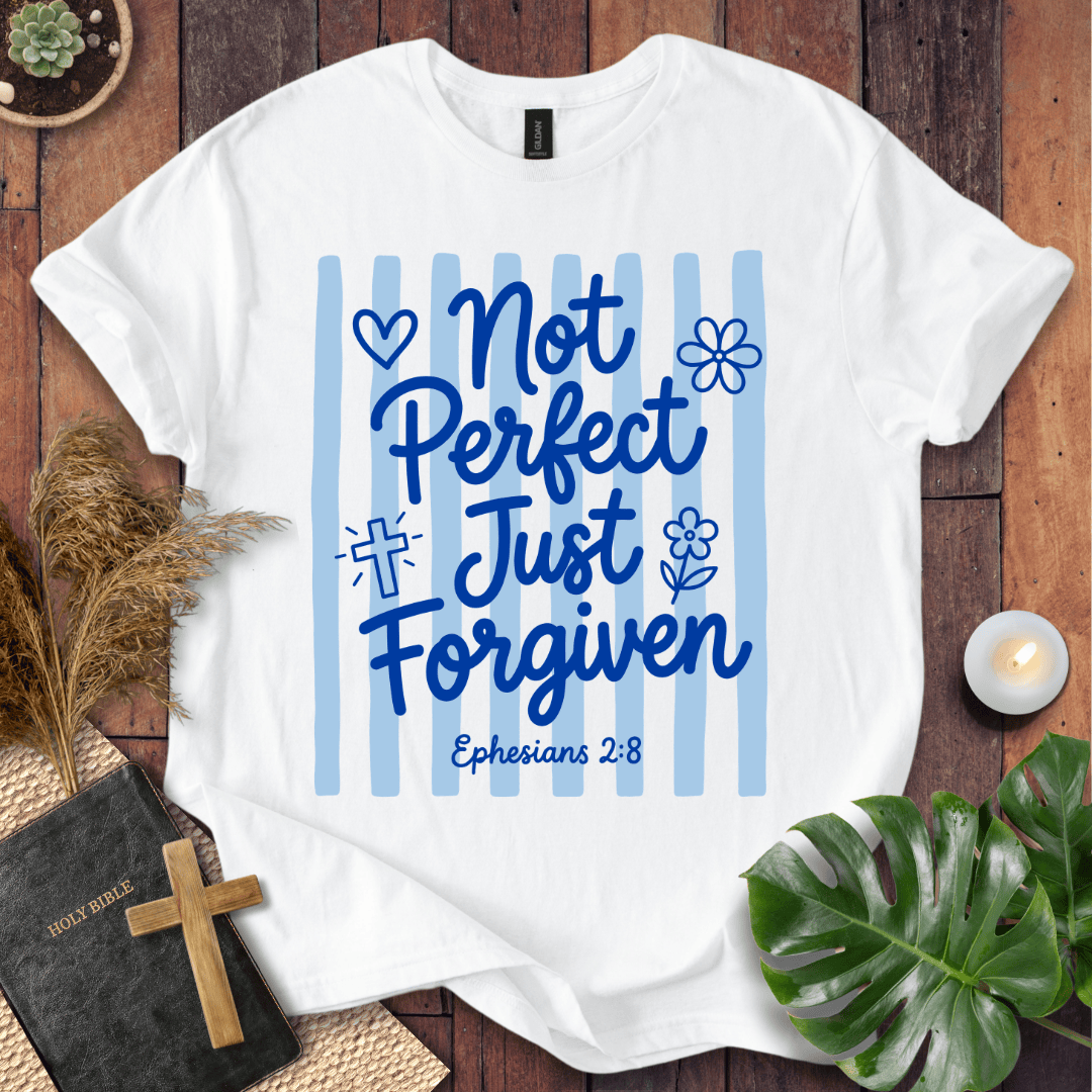 A white short-sleeve graphic T-shirt laid flat on a rustic wooden surface, surrounded by decorative props including a Bible with a wooden cross, dried grass, a small candle, green leaves, and a small plant pot. The shirt has a blue striped chest graphic with the words 'Not Perfect Just Forgiven' and 'Ephesians 2:8' in blue lettering.
