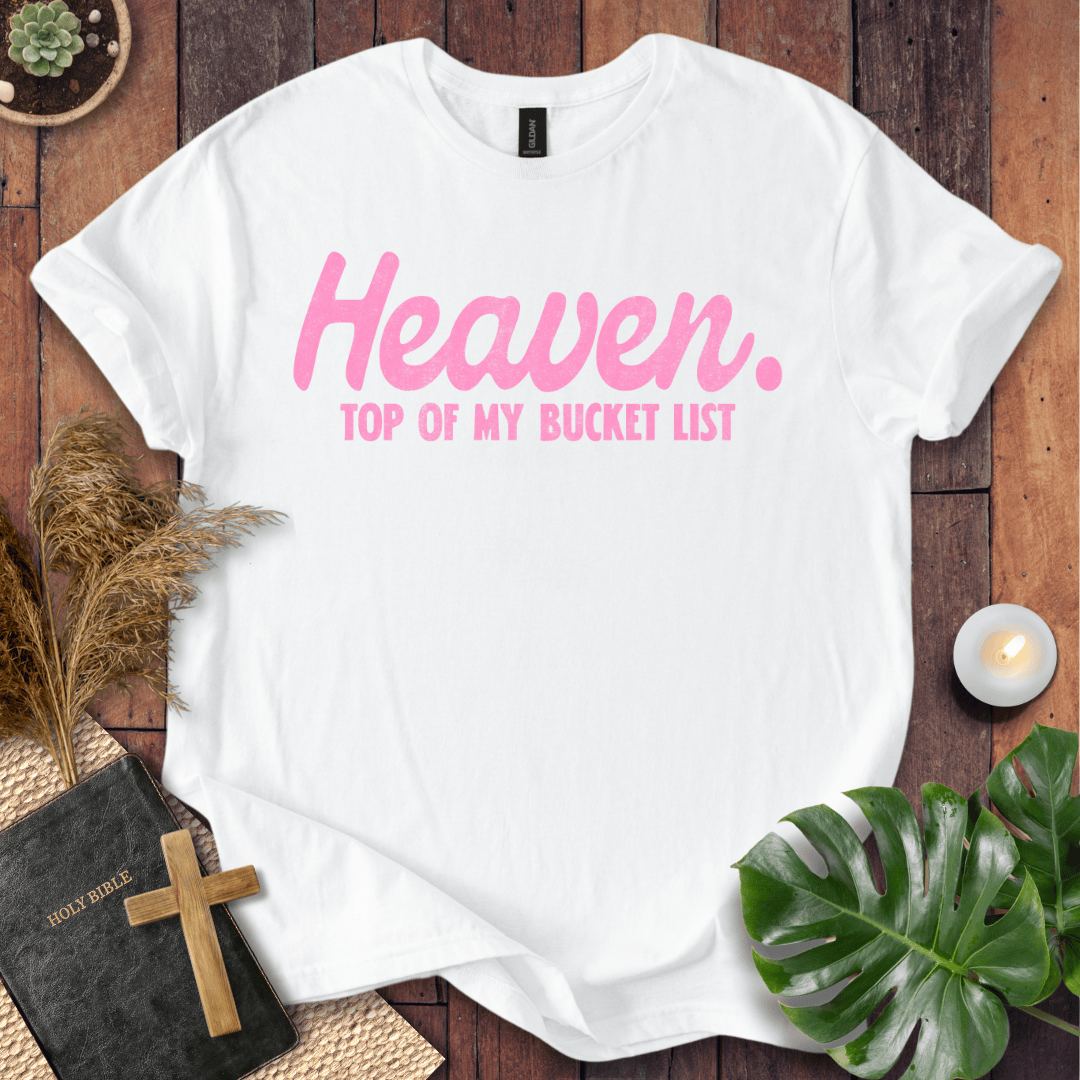 A white short-sleeve T-shirt laid flat on a rustic wooden surface, printed with the phrase 'Heaven. TOP OF MY BUCKET LIST' in pink lettering.