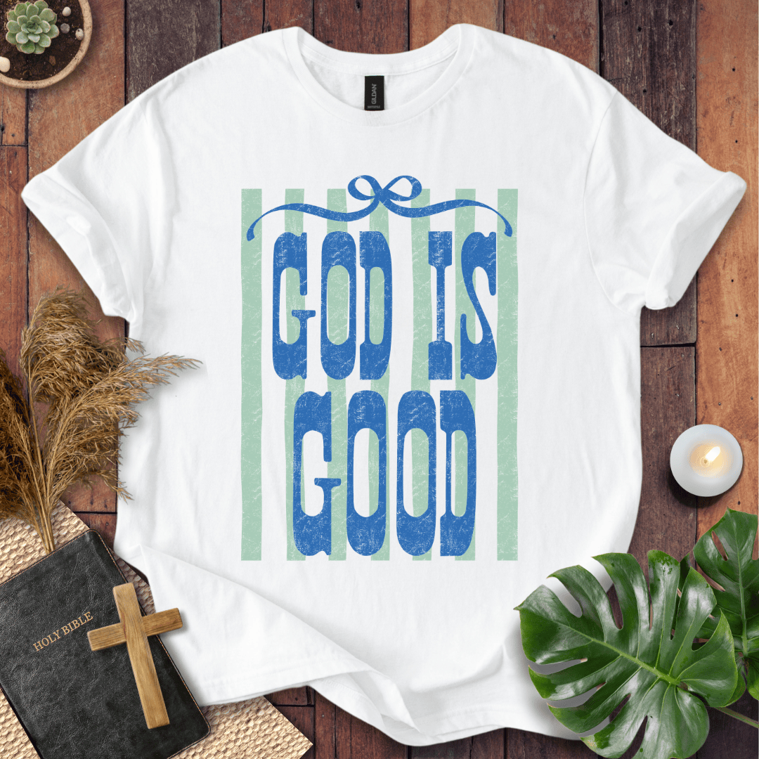 A white short-sleeve T-shirt laid flat on a rustic wooden surface, featuring the phrase 'GOD IS GOOD' in blue textured letters over pale green vertical stripes. The shirt is styled with a black Bible and wooden cross, dried grass, a lit candle, and green leaves.