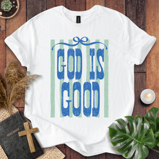 A white short-sleeve T-shirt laid flat on a rustic wooden surface, featuring the phrase 'GOD IS GOOD' in blue textured letters over pale green vertical stripes. The shirt is styled with a black Bible and wooden cross, dried grass, a lit candle, and green leaves.