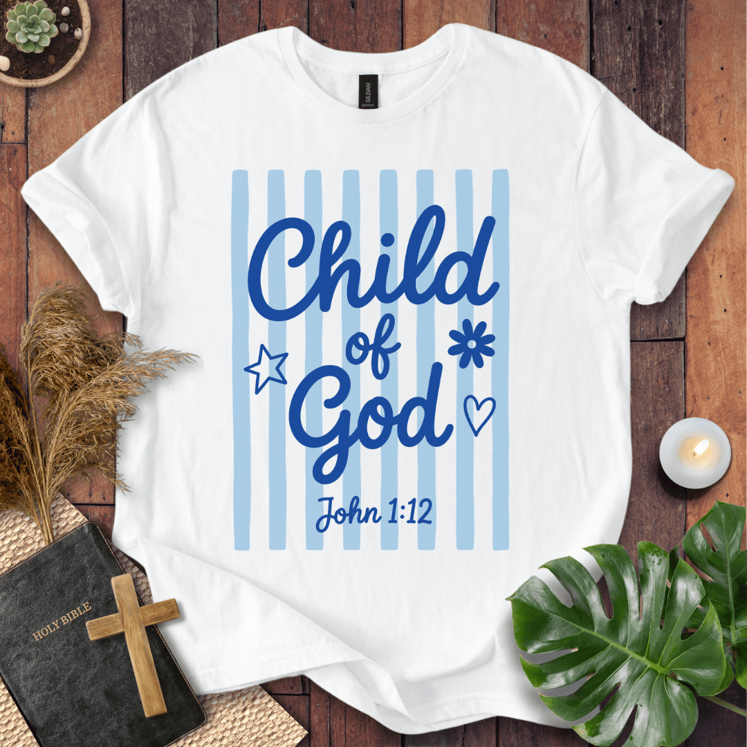 A white short-sleeve T-shirt laid flat on a rustic wooden surface, featuring the phrase 'Child of God' in blue script over light blue vertical stripes, with 'John 1:12' below. The shirt is styled with a Bible and cross, dried grass, a lit candle, and green leaves.