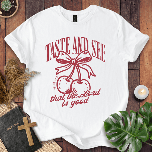 A white short-sleeve graphic T-shirt laid flat on a rustic wood surface, surrounded by decorative props including a Bible with a wooden cross, dried grass, a lit candle, green leaves, and a small plant. The shirt has a red printed design featuring the words “TASTE AND SEE” above two cherries tied with a bow, and “that the Lord is good” below.
