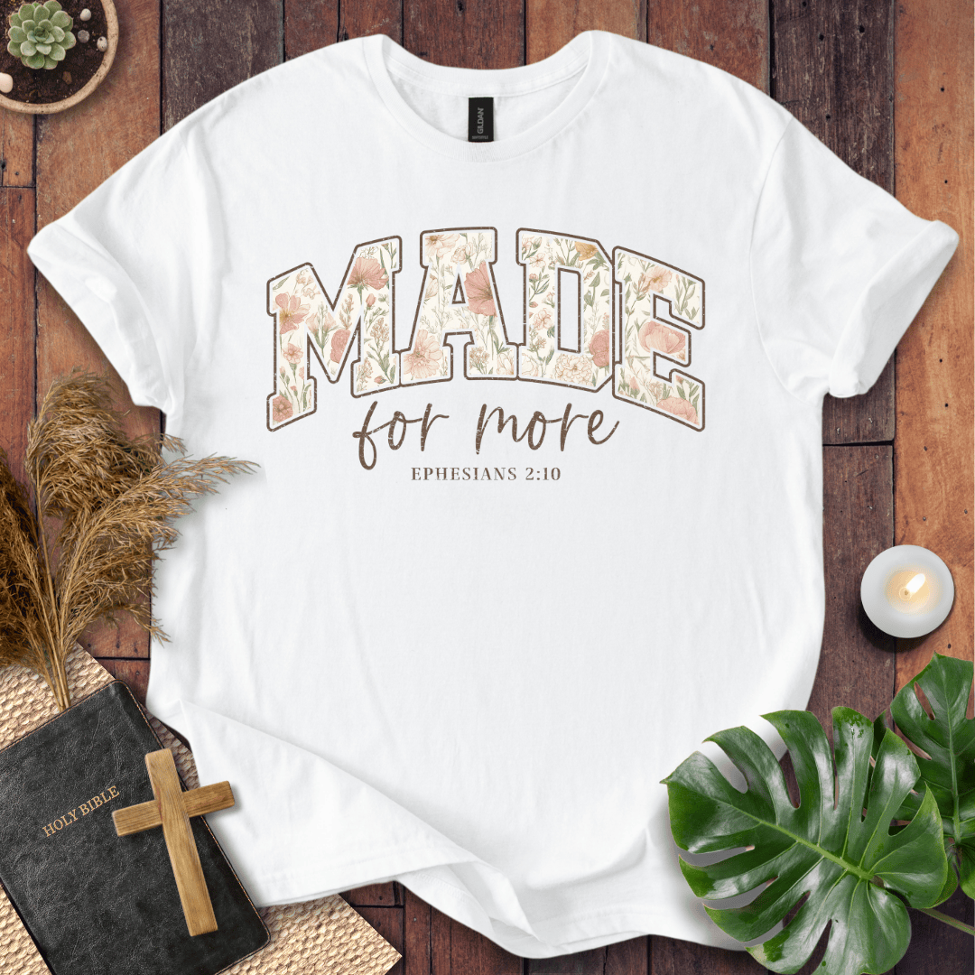 A white short-sleeve graphic T-shirt laid flat on a rustic wood surface, surrounded by decorative props including a Bible with a wooden cross, dried grass, a lit candle, green leaves, and a small plant. The shirt has a large arched 'MADE' graphic filled with a floral pattern in soft peach, beige, and muted green tones, with 'for more' in brown script and 'EPHESIANS 2:10' below in small brown text.