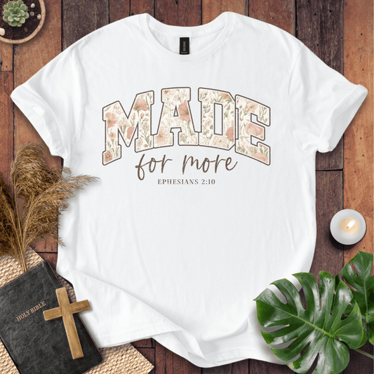 A white short-sleeve graphic T-shirt laid flat on a rustic wood surface, surrounded by decorative props including a Bible with a wooden cross, dried grass, a lit candle, green leaves, and a small plant. The shirt has a large arched 'MADE' graphic filled with a floral pattern in soft peach, beige, and muted green tones, with 'for more' in brown script and 'EPHESIANS 2:10' below in small brown text.