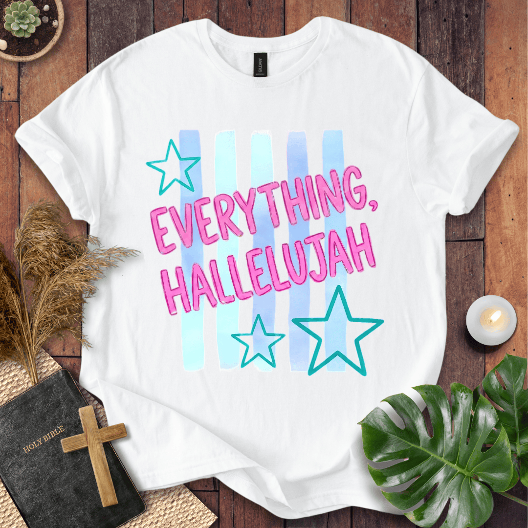 A white short-sleeve graphic T-shirt laid flat on a wooden surface, featuring the phrase 'EVERYTHING, HALLELUJAH' in pink outlined letters over pastel blue and light blue vertical stripes, with two teal outlined stars. The shirt is styled with a Bible, cross, pampas grass, green leaves, and a small candle.