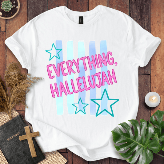 A white short-sleeve graphic T-shirt laid flat on a wooden surface, featuring the phrase 'EVERYTHING, HALLELUJAH' in pink outlined letters over pastel blue and light blue vertical stripes, with two teal outlined stars. The shirt is styled with a Bible, cross, pampas grass, green leaves, and a small candle.
