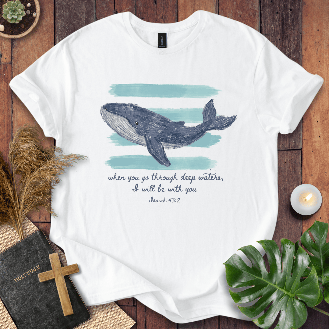 A white short-sleeve graphic T-shirt laid flat on a rustic wooden surface, featuring a watercolor-style blue-gray whale illustration above teal brushstroke bands and the printed quote, 'when you go through deep waters, I will be with you Jesus 43:2.' Decorative props including a Bible with a cross, dried grass, a candle, and green leaves surround the shirt.