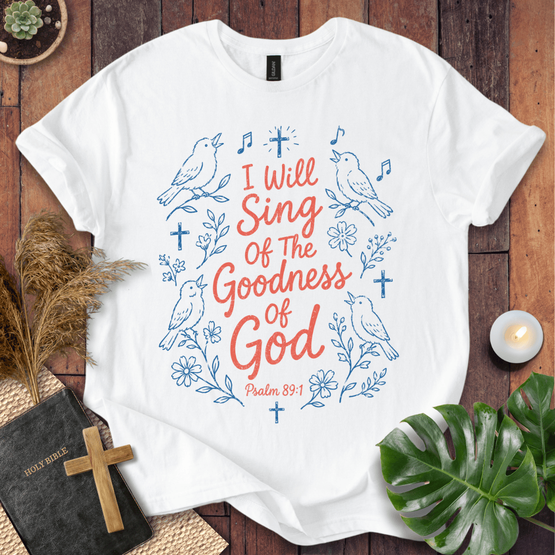 A white short-sleeve T-shirt laid flat on a rustic wooden surface, featuring blue line-art birds, flowers, and crosses with the phrase 'I Will Sing Of The Goodness Of God' and 'Psalm 89:1' in coral-red lettering. Decorative props including a book with a cross, dried grass, a candle, and green leaves surround the shirt.