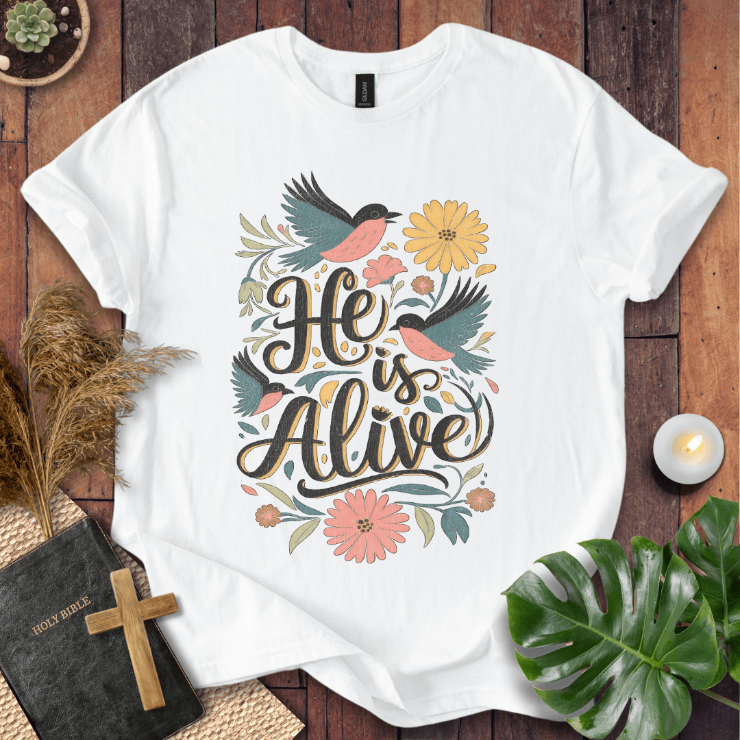 He Is Alive" Christian Easter t shirt for women with birds and flowers on a natural cream shirt, styled on a rustic wood background.