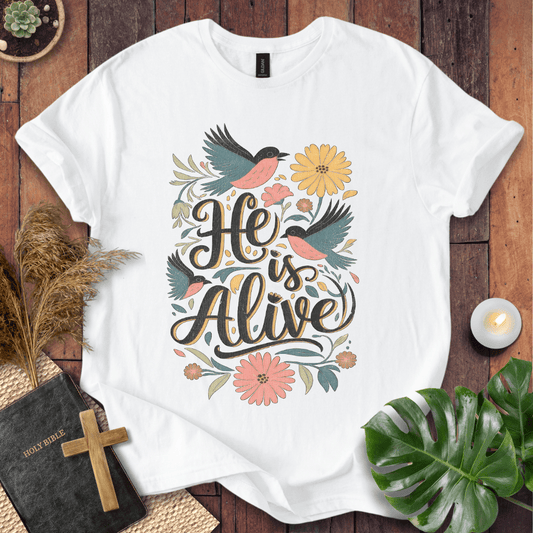 He Is Alive" Christian Easter t shirt for women with birds and flowers on a natural cream shirt, styled on a rustic wood background.
