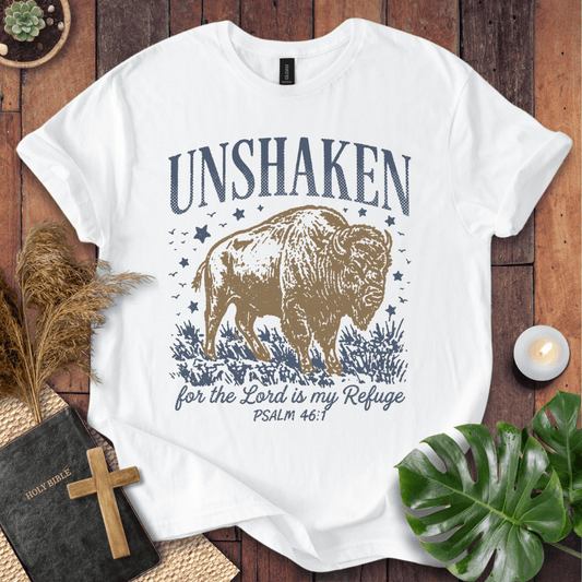 A white graphic t-shirt laid flat on a rustic wooden surface, surrounded by decorative props including a Bible with a cross, dried grass, a lit candle, green leaves, and a small plant. The shirt features a graphic of a bison with the text 'UNSHAKEN for the Lord is my Refuge PSALM 46:1' in navy blue and tan.