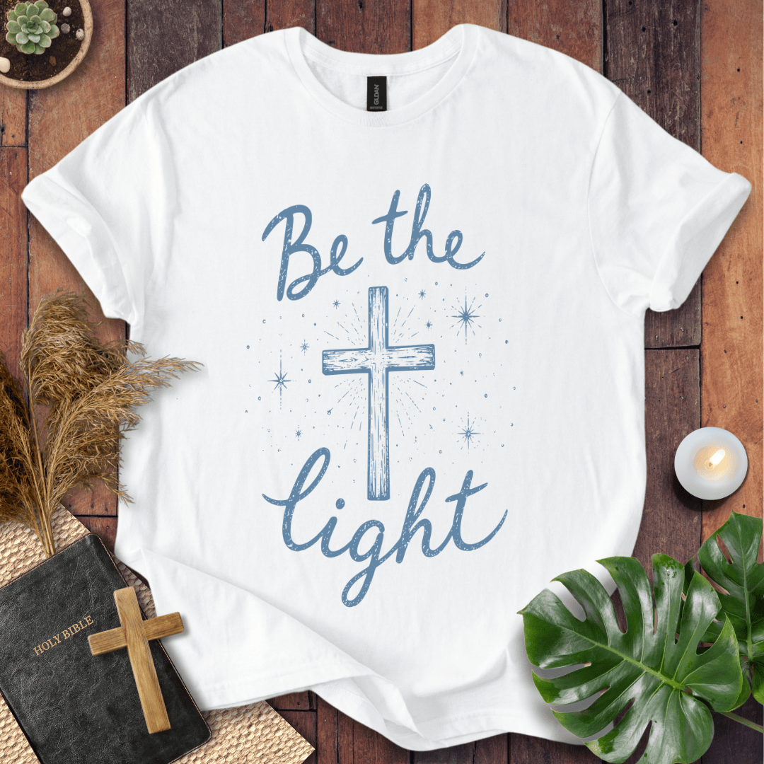 Christian t shirt for women with blue "Be the Light" text, a central cross, and starry accents on a wooden background.